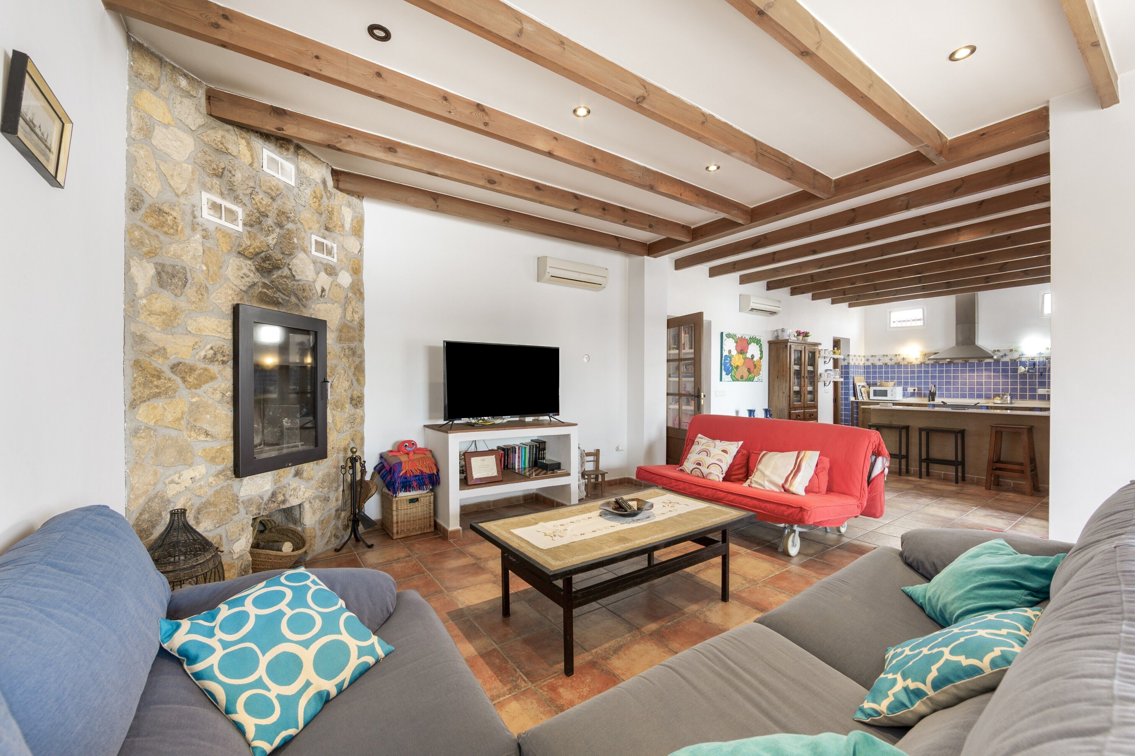 A living room with a stone wall, wooden beams, a television, and a kitchen in the background.