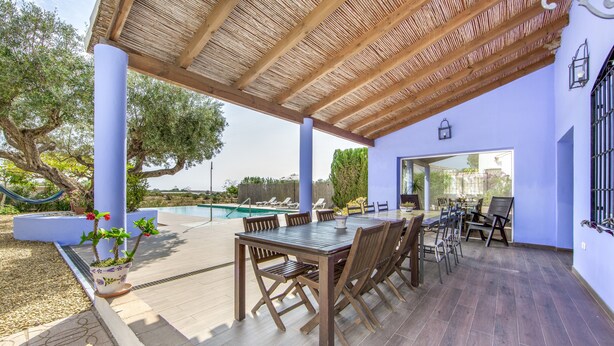 An outdoor patio with a wooden table and chairs, a pool, and a covered area with a thatched roof. - My Grandparents’ Country House (Los Gallardos)