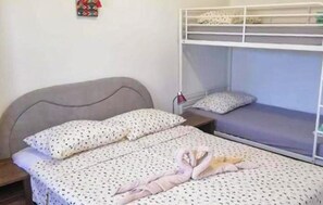 Ein Etagenbett mit grauem Kopfteil, ein Einzelbett mit einem gemusterten Bettüberwurf und ein kleiner Nachttisch mit Lampe.
