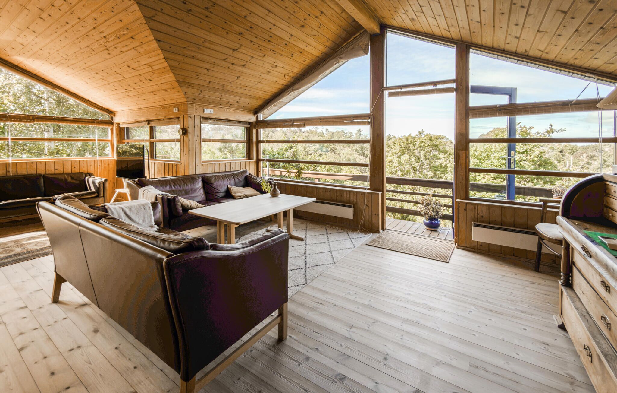 A wooden cabin interior with a sofa, table, and large windows offering a view of the outdoors.