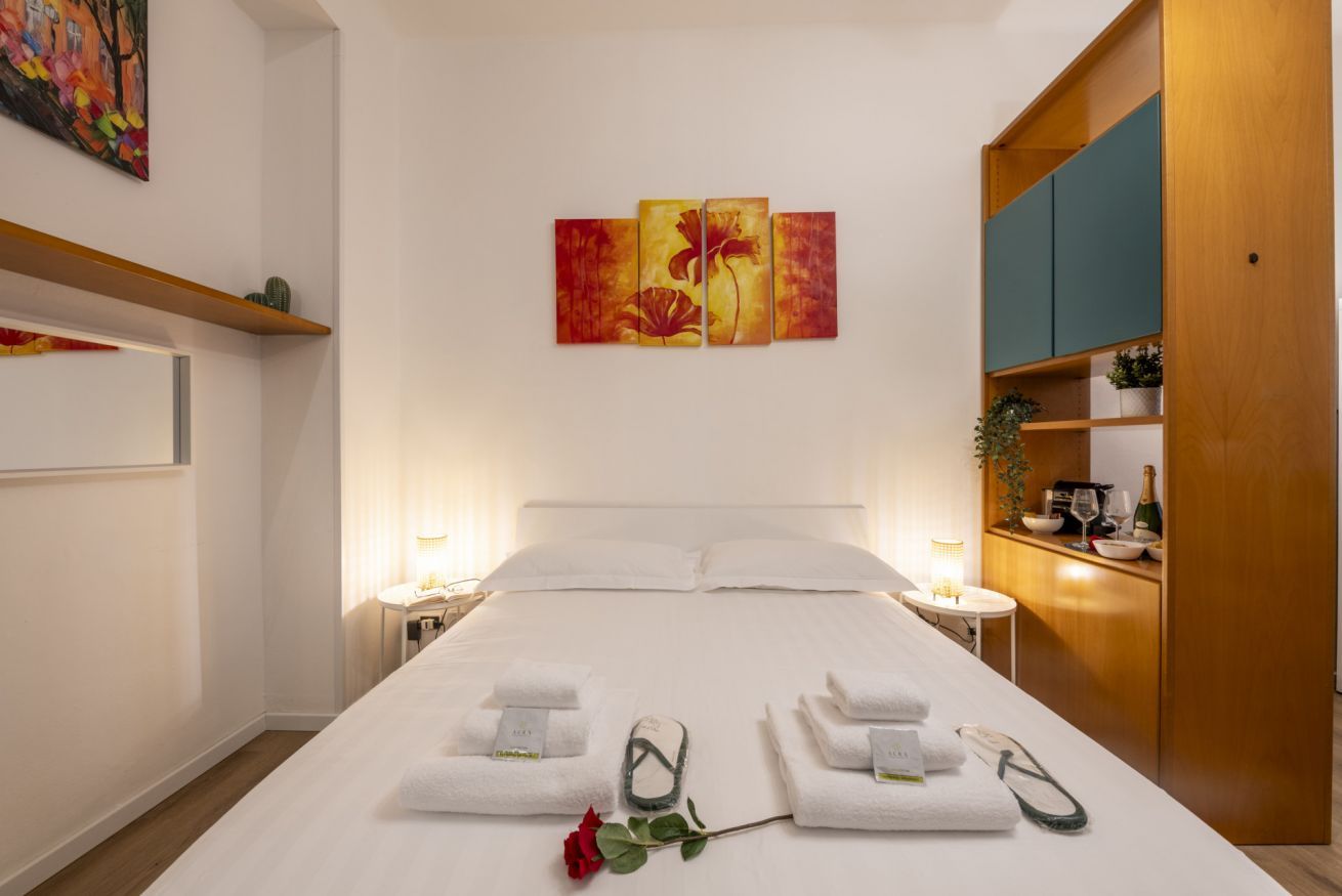 A hotel room with a large bed, a painting on the wall, a wooden cabinet, and a bedside table with a lamp.