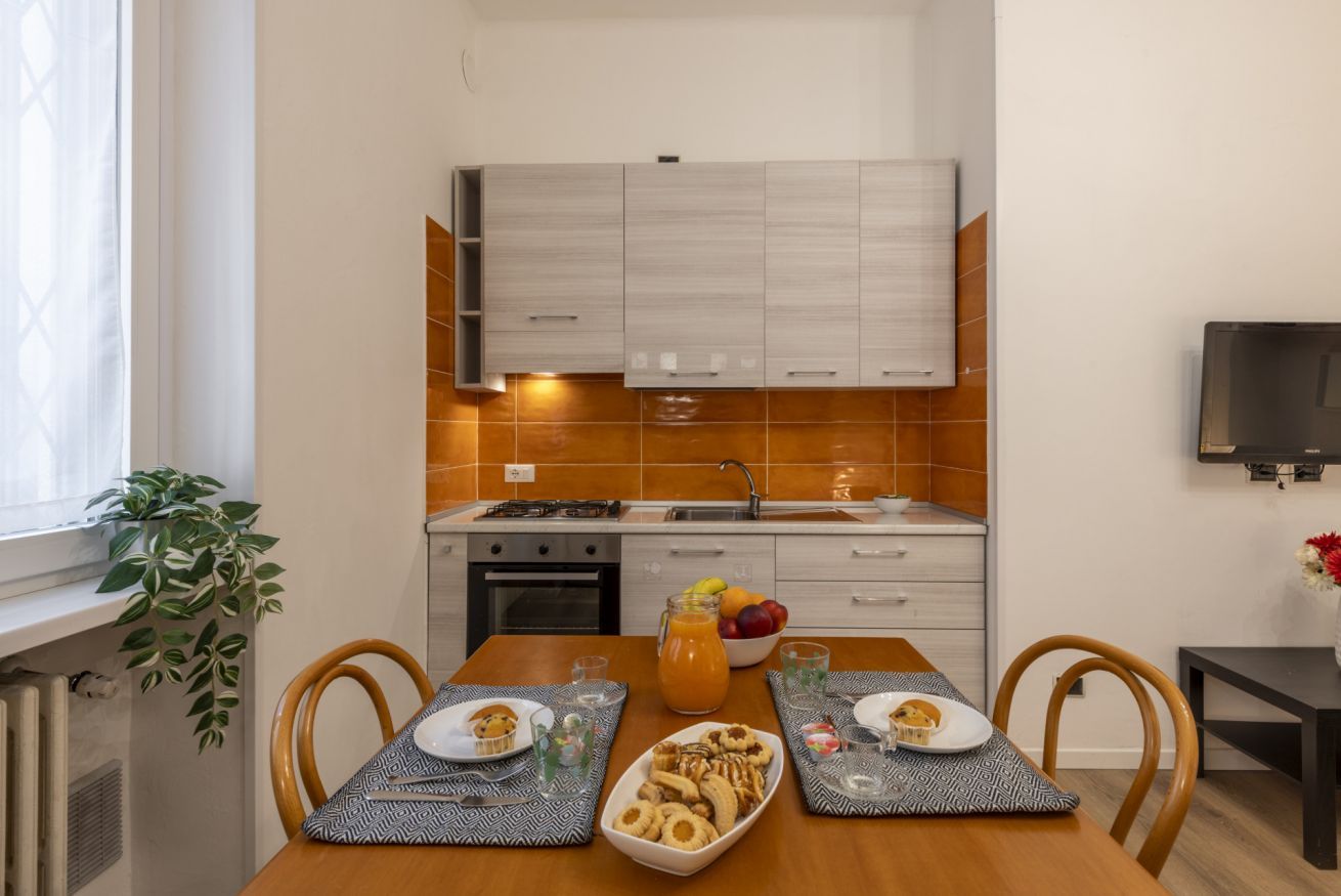 A compact kitchen with modern appliances, a dining table set with plates and glasses, and a potted plant by the window.