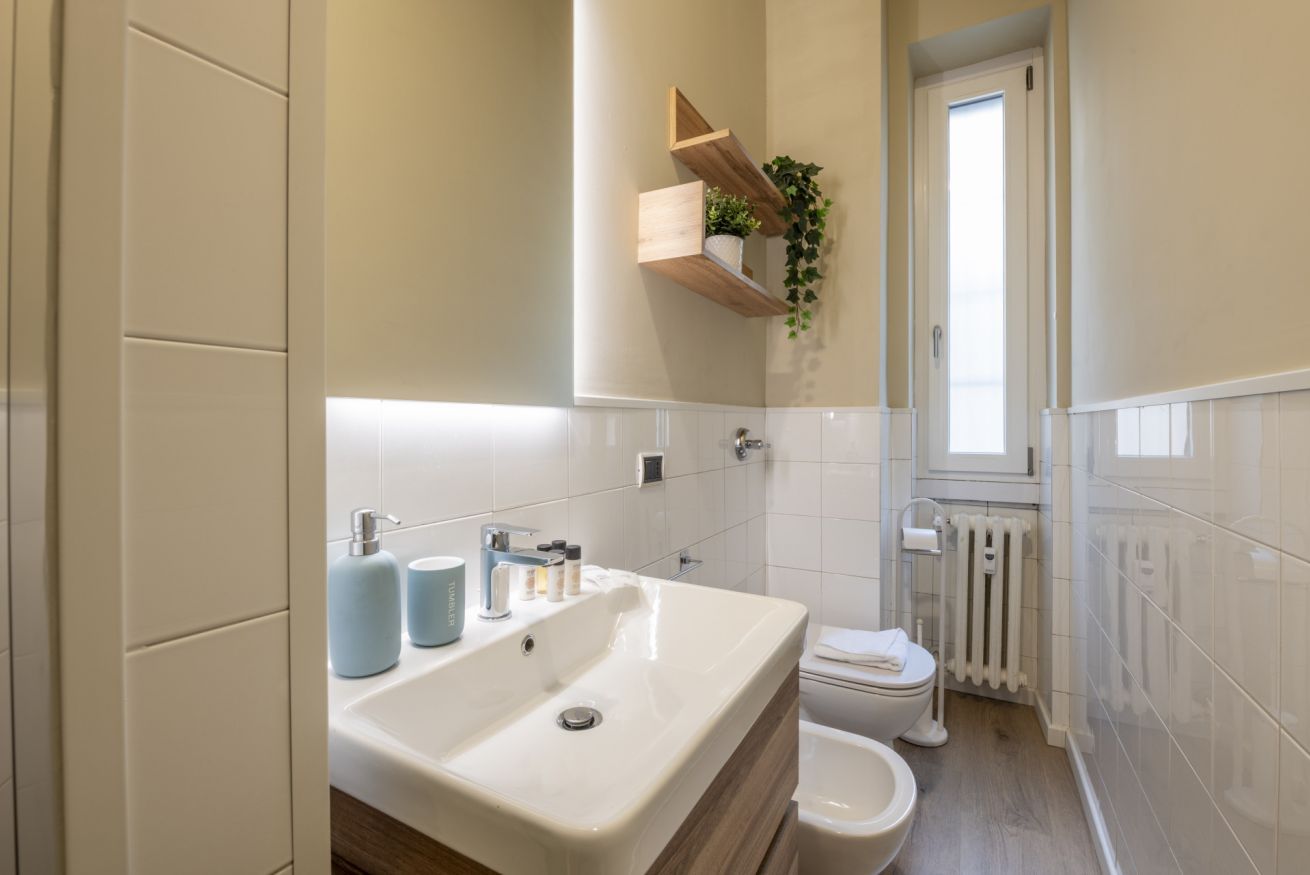 Une salle de bain avec un lavabo, des toilettes et un radiateur blancs, des étagères en bois avec une plante, et une fenêtre.
