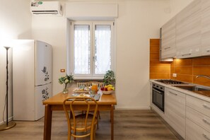 Une cuisine compacte avec une table à manger préparée pour le petit-déjeuner, un réfrigérateur et une fenêtre avec des stores.