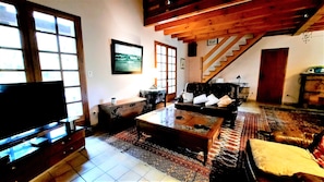 A living room with a sofa, a coffee table, a TV, a staircase, and a patterned rug. - Charming country house between the forest and the ocean - Contis (Lit-et-Mixe)