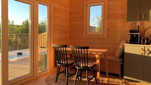 A wooden cabin interior with a dining table, chairs, a kitchen area, and large windows. - Log cabin with hot tub and views (Bondemölla)