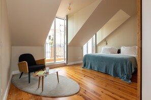 Um quarto com cama, uma cadeira, uma mesinha e uma janela grande.