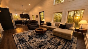 A cozy living room with a sofa, armchair, and coffee table, a kitchen in the background, and large windows with a view of trees. - Romantic Retreat with Hot Tub & Fire Pit in Broken Bow, Oklahoma (Broken Bow)