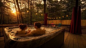 Ein Whirlpool mit zwei Personen, umgeben von einer Holzterrasse und Bäumen, mit Lichterketten im Hintergrund.