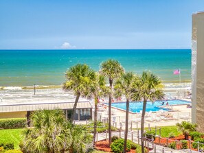 A pool area with a view of the ocean, palm trees, and a flag. - Wake Up to Ocean Views | Castle Reef 411 (New Smyrna Beach)
