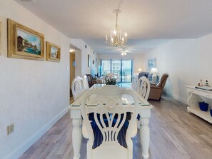 A dining area with a white table and chairs, a chandelier, and a view to the outside. - Wake Up to Ocean Views | Castle Reef 411 (New Smyrna Beach)