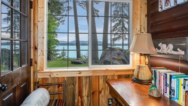 Interno di una baita di legno con un'ampia finestra che si affaccia su un lago e alberi, una lampada e libri su un tavolo di legno.