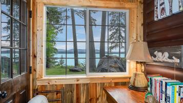 A wooden cabin interior with a large window overlooking a lake and trees, a lamp, and books on a wooden table.