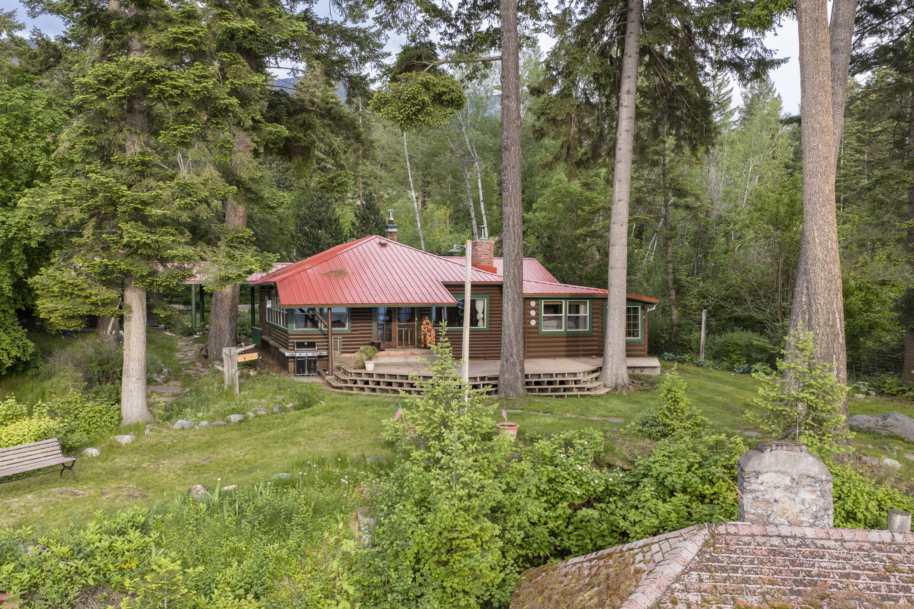 A cabin with a red roof, surrounded by dense forest, with a porch and a stone wall.