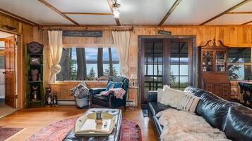 A cozy cabin interior with a wooden interior, a fireplace, a leather sofa, a reading chair, and a view of a lake.