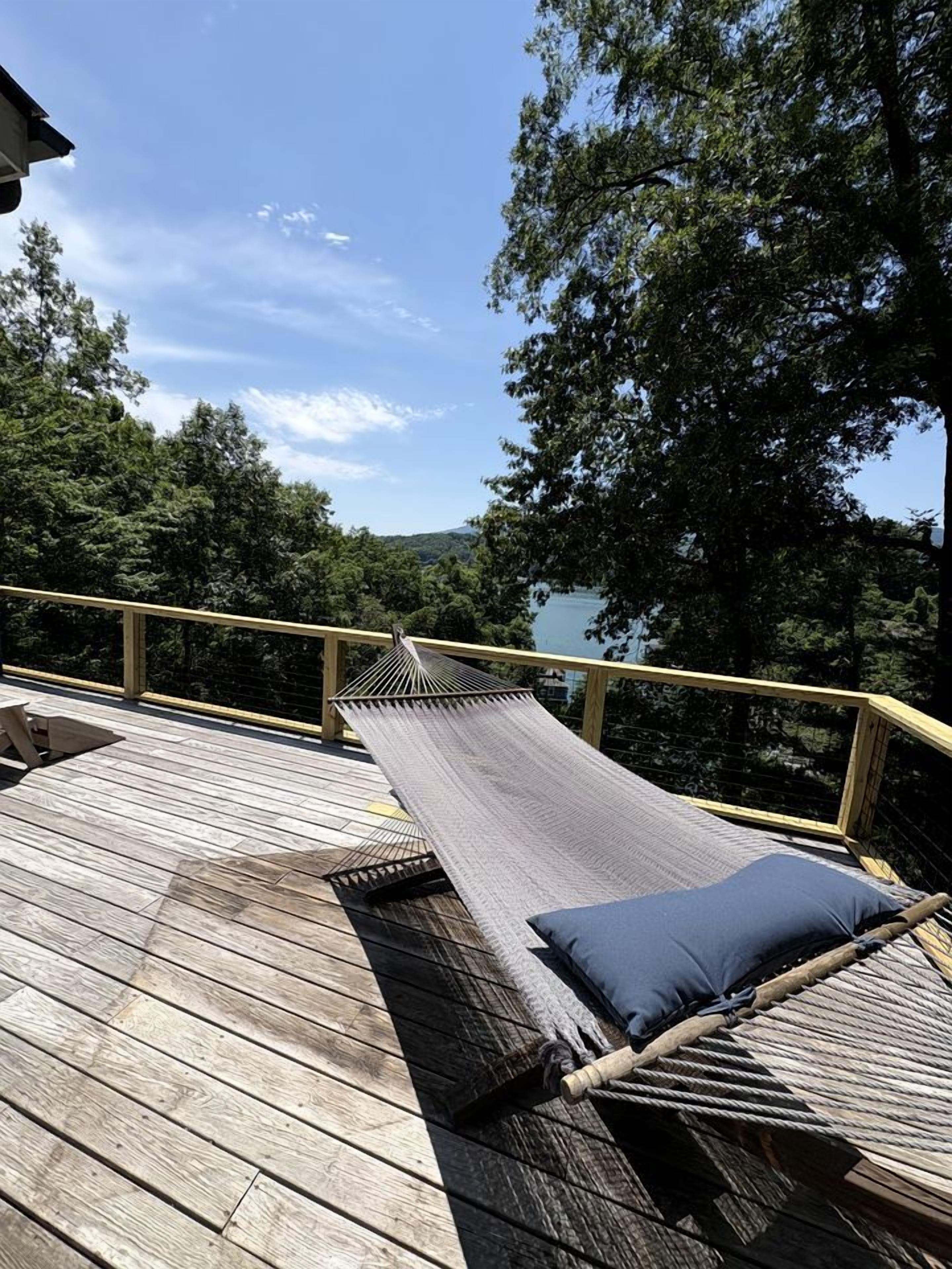 Eine Holzterrasse mit einer Hängematte und einem Kissen, mit Blick auf bewaldetes Gelände und ein Gewässer.
