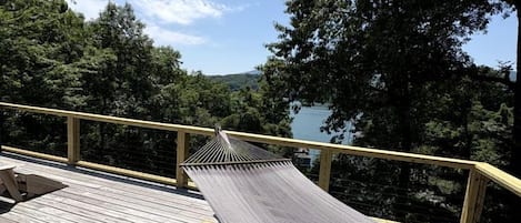 Eine Holzterrasse mit einer Hängematte und einem Kissen, mit Blick auf bewaldetes Gelände und ein Gewässer.