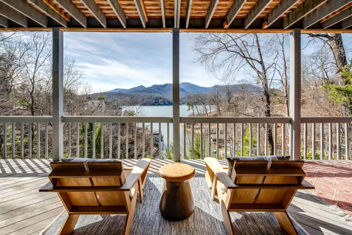 A covered outdoor deck with two wooden chairs and a round table, overlooking a lake and mountains.