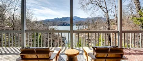 A covered outdoor deck with two wooden chairs and a round table, overlooking a lake and mountains.