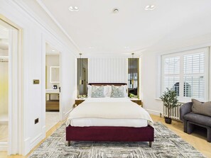 A spacious bedroom with a large bed, bedside tables, a dresser, a mirror, and a window with blinds. - Decadent and Exquisite Holland Park Duplex (Greater London)