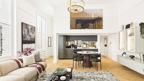 A spacious living area with a high ceiling, a dining table with chairs, a kitchen in the background, and a large window with blinds. - Decadent and Exquisite Holland Park Duplex (Greater London)