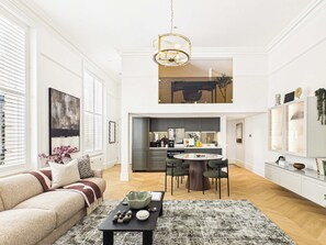 A spacious living area with a high ceiling, a dining table with chairs, a kitchen in the background, and a large window with blinds. - Decadent and Exquisite Holland Park Duplex (Greater London)