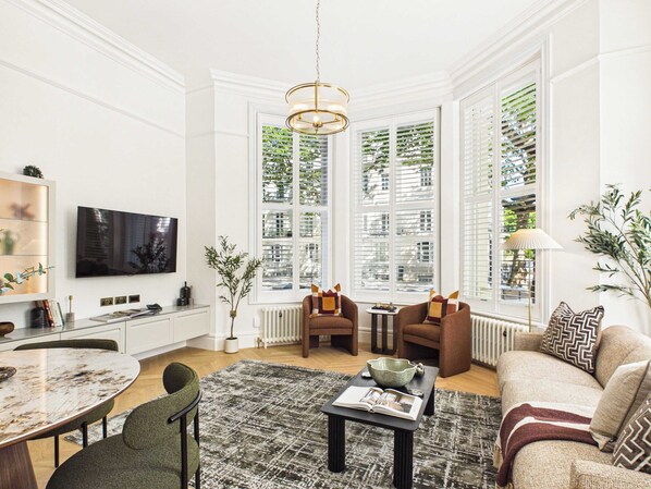 A modern living room with a dining area, large windows with blinds, a television, and a chandelier. - Decadent and Exquisite Holland Park Duplex (Greater London)