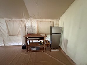Una habitación con toldo, una mesa de madera, un ventilador, un refrigerador y un recipiente de plástico.