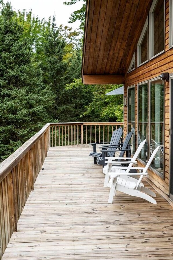 Terraza de madera con sillas Adirondack, una puerta de vidrio y vista a un bosque denso.