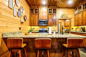 Une cuisine avec des meubles en bois, un comptoir en granit, un four à micro-ondes et une cuisinière. Il y a trois tabourets de bar en bois.