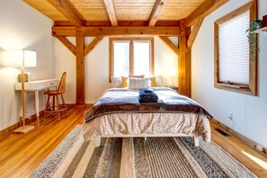 Een slaapkamer met een houten plafond, een bed met een beddengoed met dessin, een bureau met stoel, en een raam met jaloezieën.