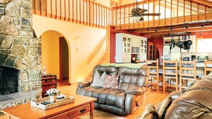 Ein gemütliches Wohnzimmer mit Kamin aus Stein, Ledersofa, Holztisch und einer Küche im Hintergrund.