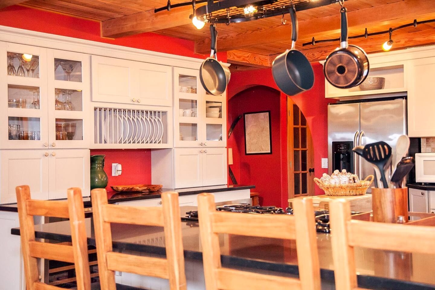 A kitchen with white cabinets, a red accent wall, hanging pots, and a central island with chairs.