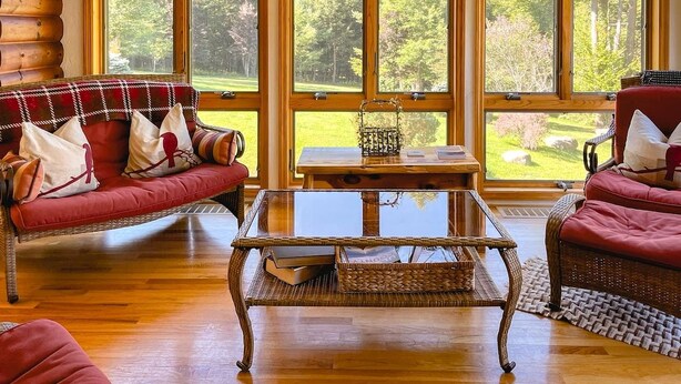 Ein gemütliches Wohnzimmer mit einem roten Sofa, einem Korbtisch und Blick auf einen Wald durch große Fenster.