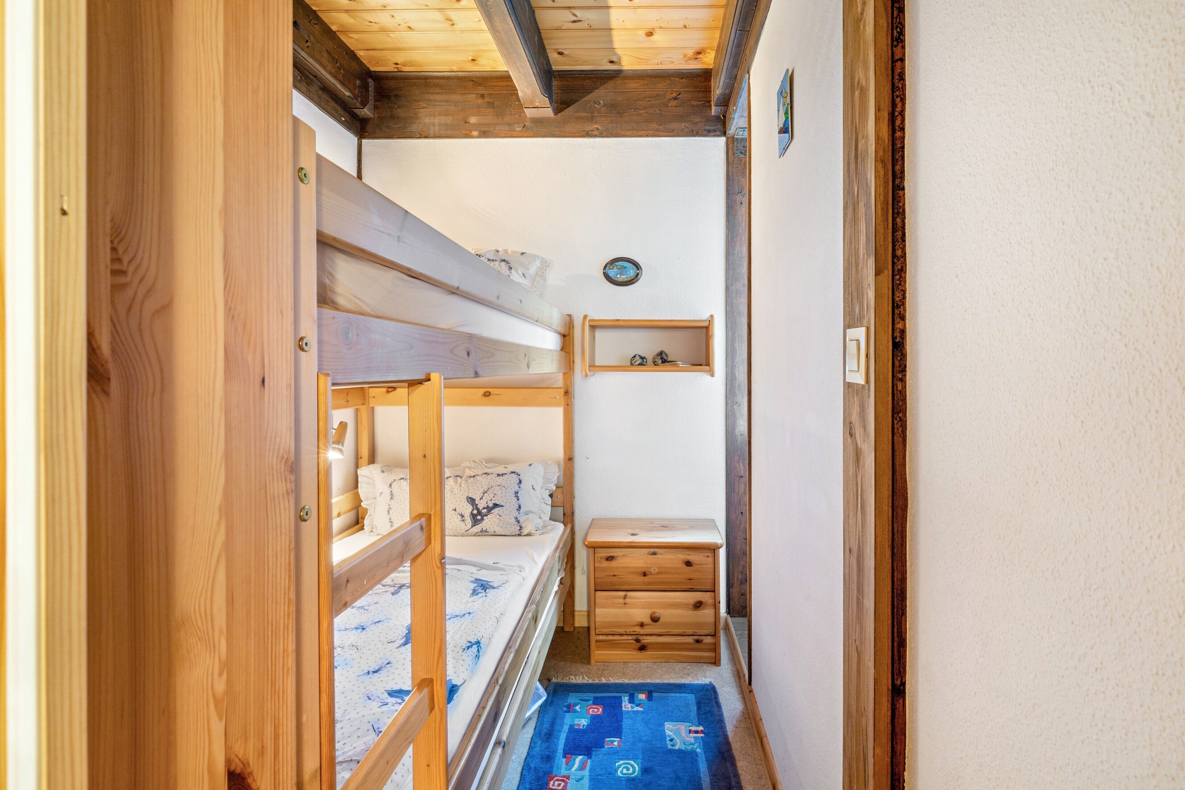 A narrow room with a bunk bed, a small wooden dresser, and a shelf with items on the wall.