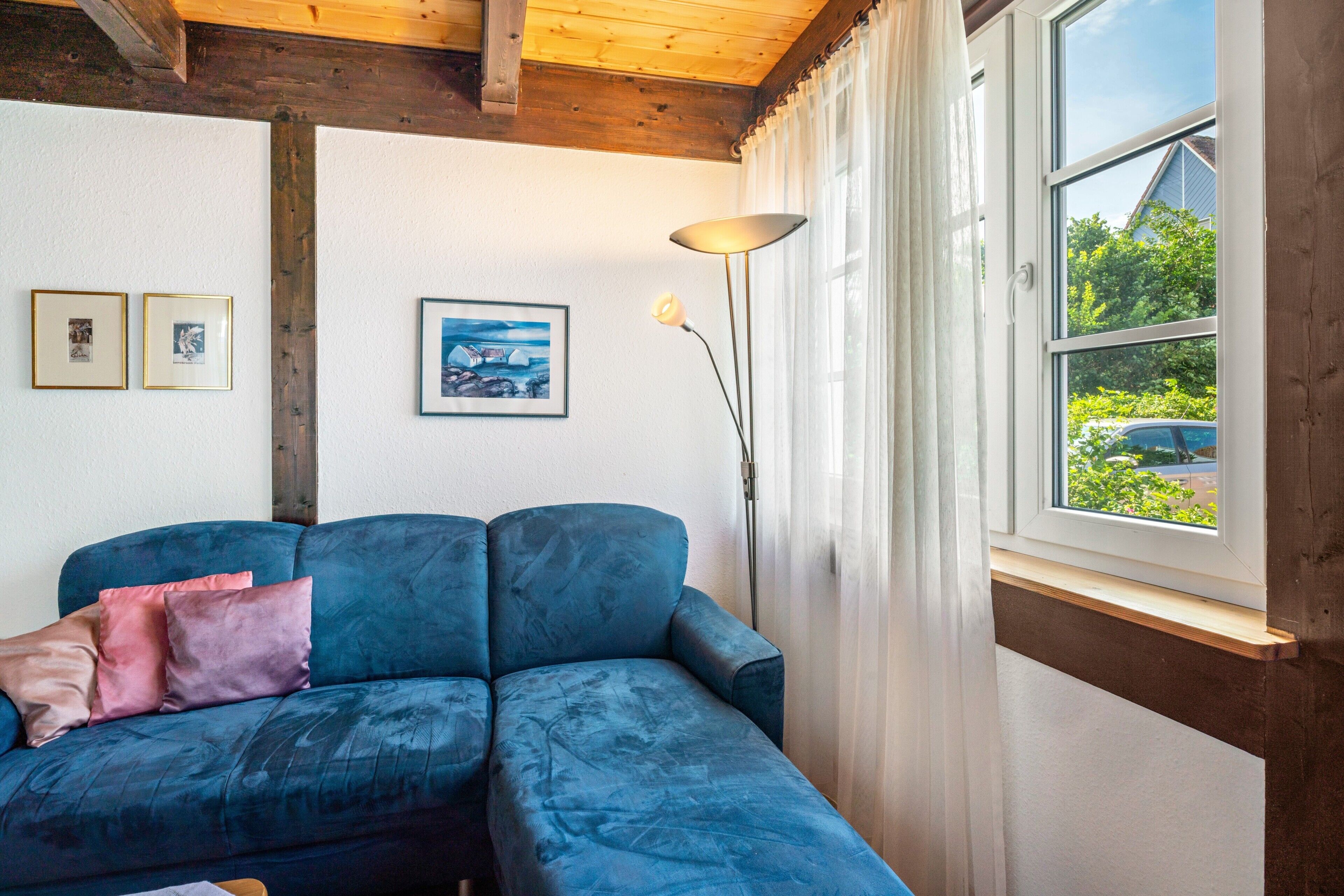 A room with a blue sofa, a lamp, and a window with a view of greenery and a building.