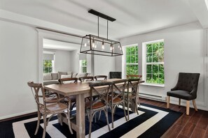 Un espace repas comprenant une table en bois, des chaises, un luminaire suspendu et de grandes fenêtres donnant sur un paysage verdoyant.