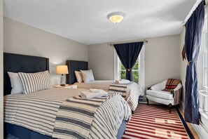 Ein Hotelzimmer mit zwei Betten, einem Nachttisch, einer Lampe, einem Sessel und einem Fenster mit Vorhängen.