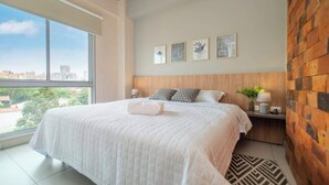 A bedroom with a large window, a bed with a white quilt, a wooden headboard, and three framed pictures on the wall.