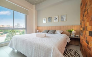 Ein Schlafzimmer mit einem großen Fenster, einem Bett mit weißer Bettdecke, einem Holz-Kopfteil und drei gerahmten Bildern an der Wand.
