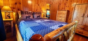 Ein Zimmer in einer Holzhütte mit einem Bett, einem Schminktisch und einem Nachttisch mit Lampe.