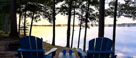 Two Adirondack chairs facing a lake with trees in the background.
