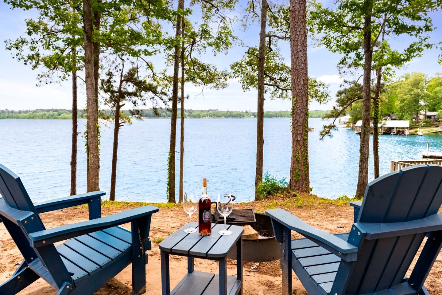 Vista de un lago con dos sillas Adirondack, una mesita y una botella de vino.