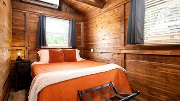 Een slaapkamer in een houten hutje, met een bed, nachtkastje, airconditioning en een raam met gordijnen.
