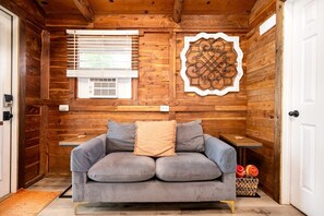 Une pièce chaleureuse aux murs en bois, avec un canapé gris agrémenté de coussins, un élément décoratif en métal et une fenêtre équipée de stores.