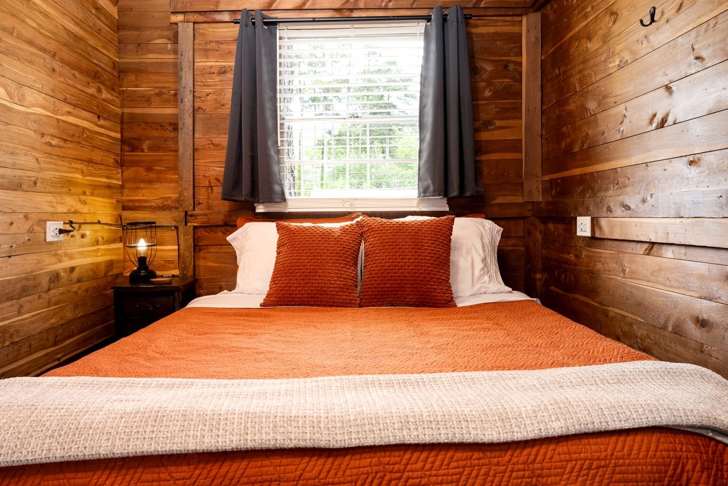 A wooden cabin-style room with a bed, a nightstand with a lamp, and a window with curtains.