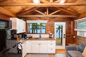 Cocina tipo cabaña con techo de madera, armarios blancos, refrigerador de acero inoxidable y vista a un lago a través de ventanas amplias.