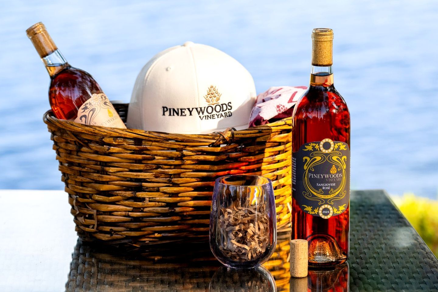 A wicker basket containing a white cap, two bottles of Piney Woods Vineyards Sangiovese, and a glass with wood shavings.