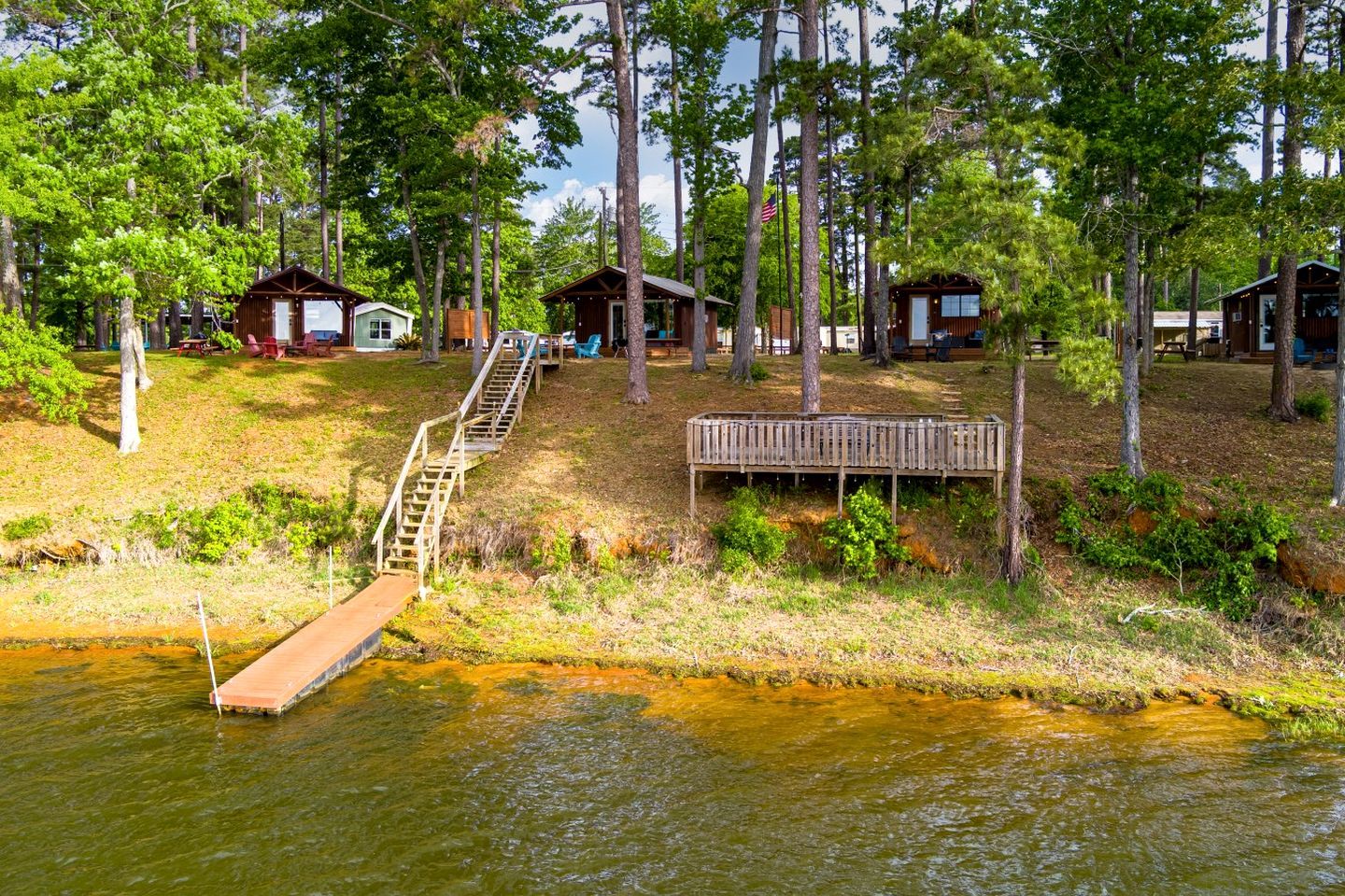 A lakeside cabin area with wooden docks, a wooden staircase, and several cabins surrounded by trees.