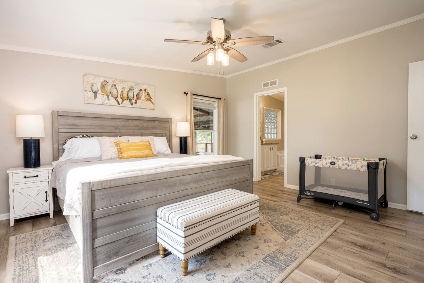 A bedroom with a bed, bedside tables, a bench, a ceiling fan, a painting, and a crib.